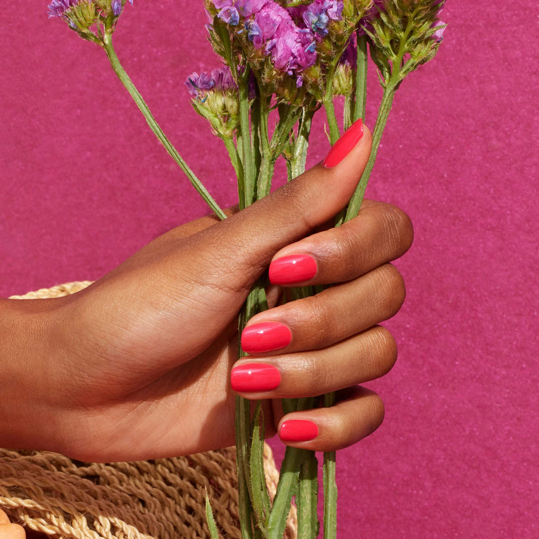 Hand mit korallrosa manikürten Nägeln hält lila Wildblumen vor einem leuchtend rosa Hintergrund und präsentiert Manucurist Bougainvillea Nagellack