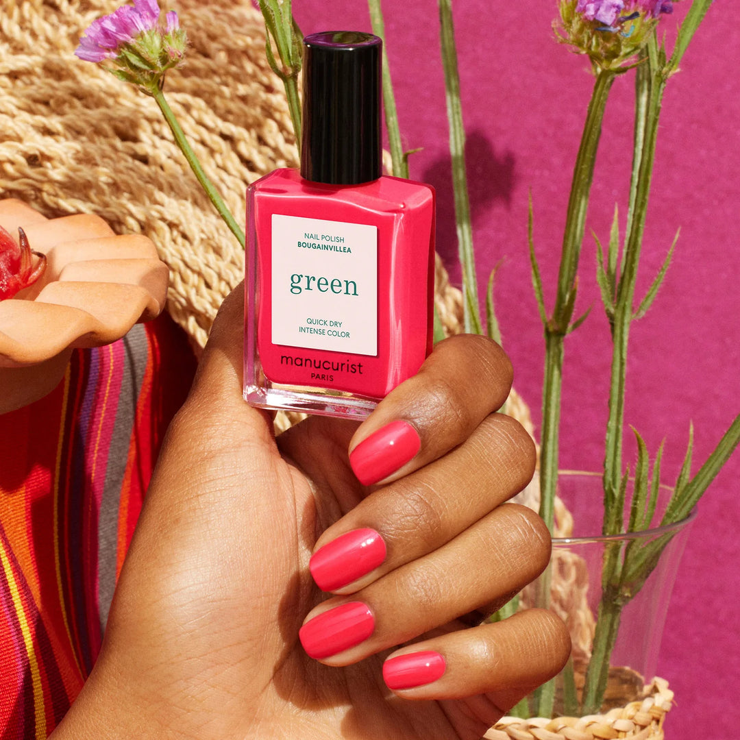 Hand holding Manucurist Paris Green nail polish in Bougainvillea shade with matching coral-pink manicure, styled with purple flowers and straw hat against pink background
