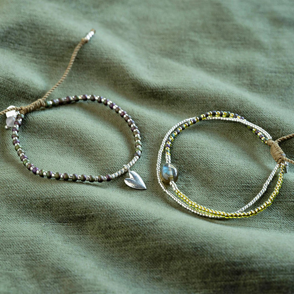 Hoping Rose Quartz Bracelet - Armband neben weiterem Schmuckstück auf dunkelgrünem Hintergrund