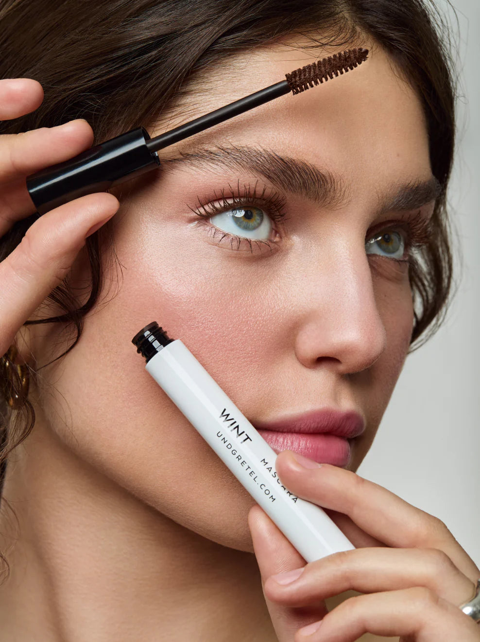 Model trägt braune Mascara auf und hält die UND GRETEL WINT Mascara in der Hand