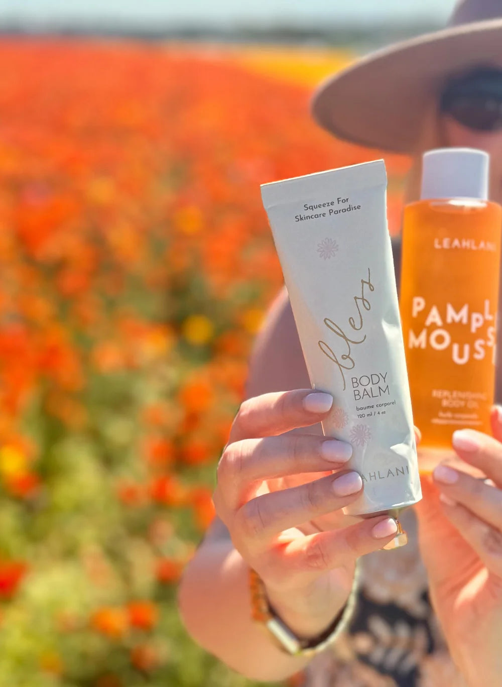 Bless Body Balm von Leahlani - mit weiterem Produkt, gehalten von Model vor Blumenfeld